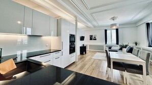 Een moderne keuken met witte kasten, een zwart aanrechtblad en ingebouwde apparatuur. Er is een eethoek met een witte tafel en stoelen, en een groot raam met gordijnen.