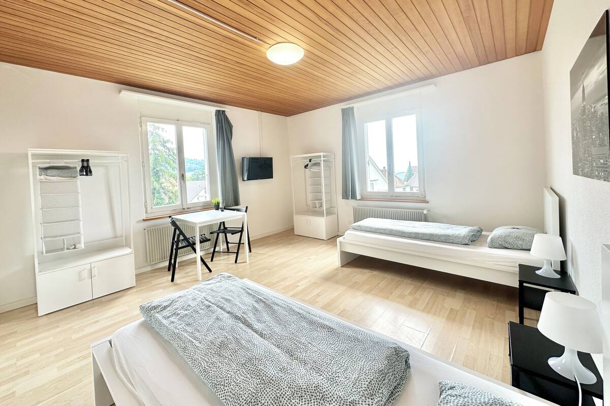 Ein modernes, helles Zimmer mit Holzboden, einem Bett, einem Schreibtisch mit Stuhl, einem Heizkörper und einem Fenster mit Blick auf Gebäude.