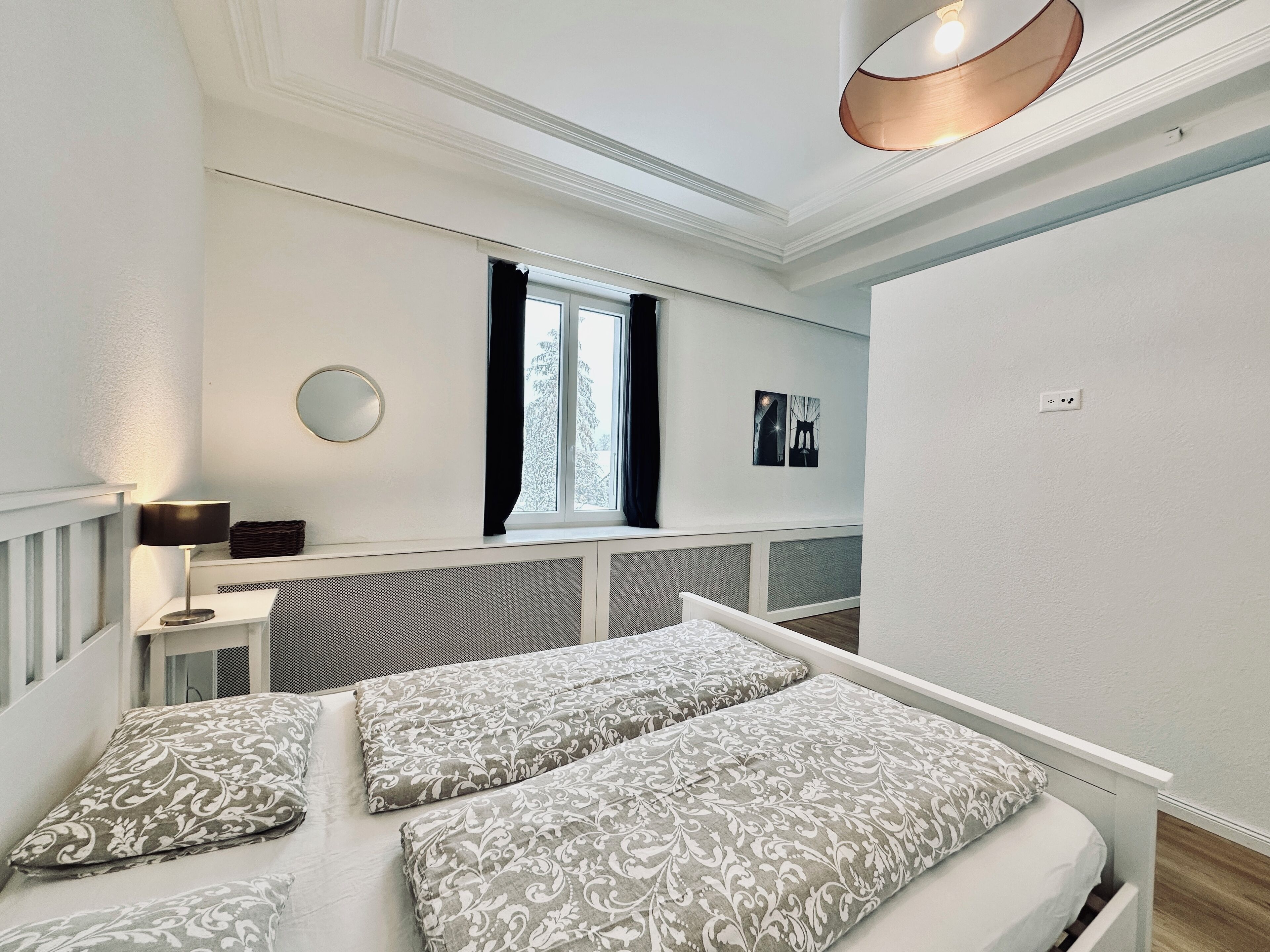 A bedroom with a bed, a bedside table, a lamp, a window with curtains, and a wall-mounted light fixture.