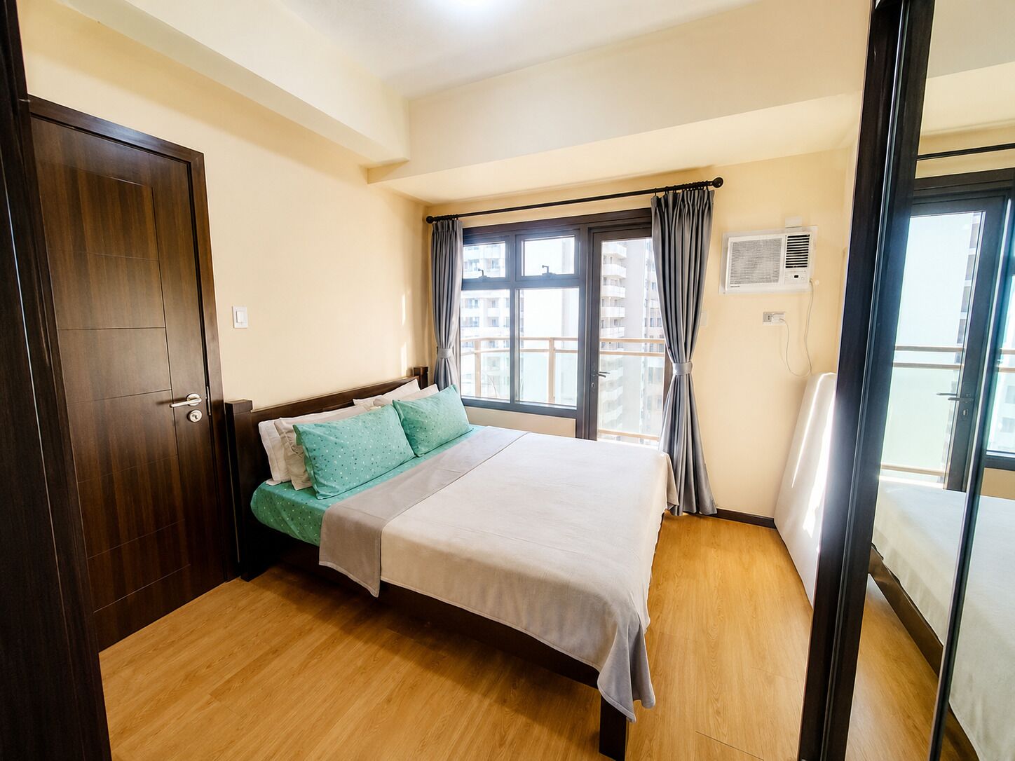 A hotel room with a bed, a window with curtains, a door, and a view of buildings outside.