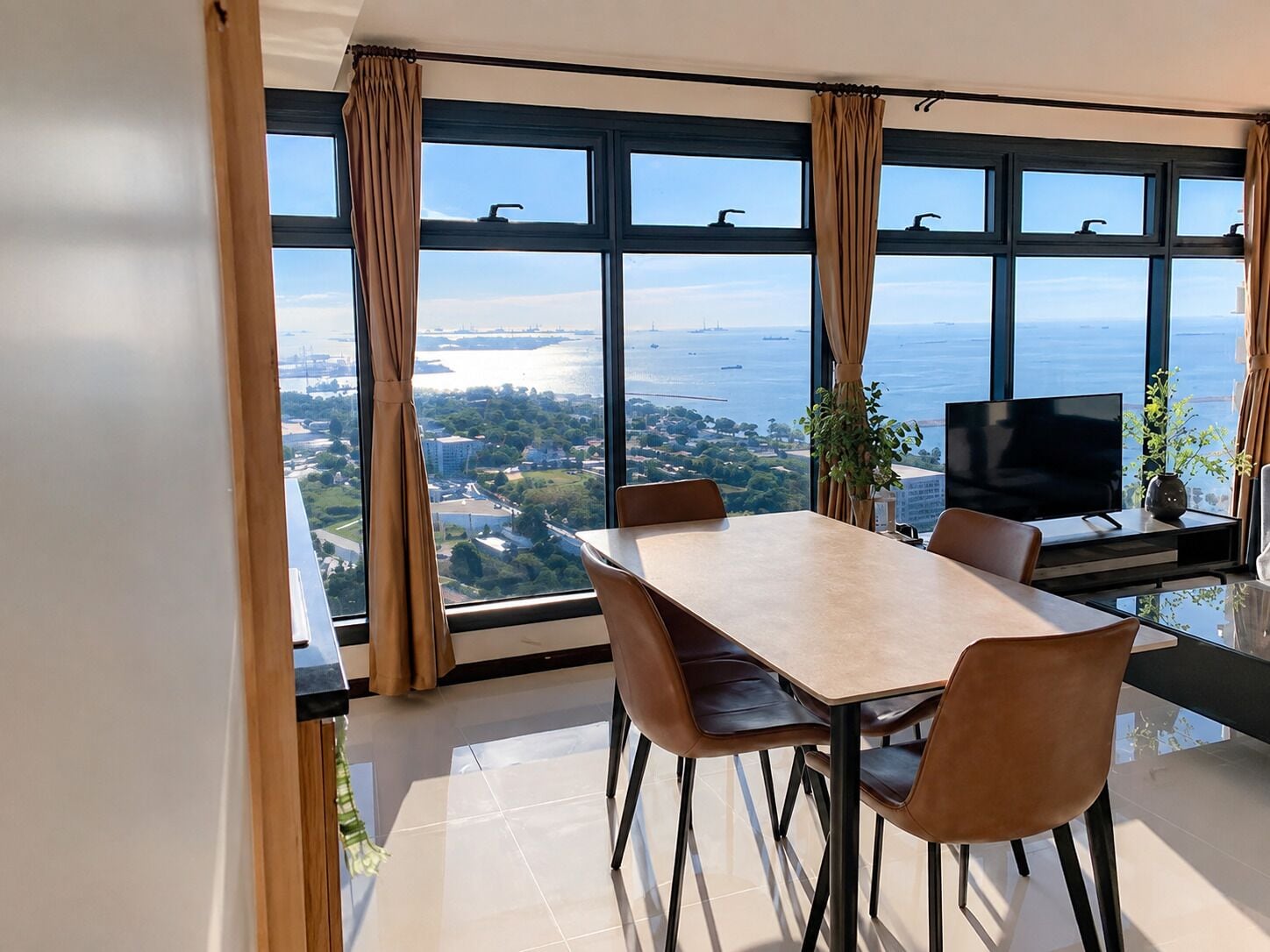 A modern dining area with a large table and chairs, a flat-screen TV, and a view of a body of water and cityscape through large windows.