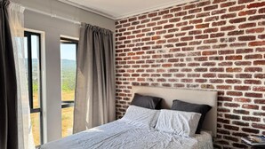 Un dormitorio con pared de ladrillo, cama con colcha estampada y vista a un paisaje por la ventana.