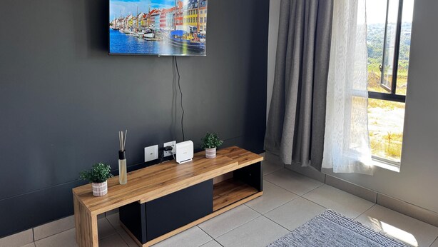 Una sala de estar moderna con un televisor de pantalla plana montado en una pared oscura, una consola de madera con dos plantas en macetas y un ventanal con vistas al exterior.