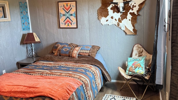 Un dormitorio con cama, ventilador de techo, un tapiz de cuero en la pared, alfombra estampada y una silla con cojín.