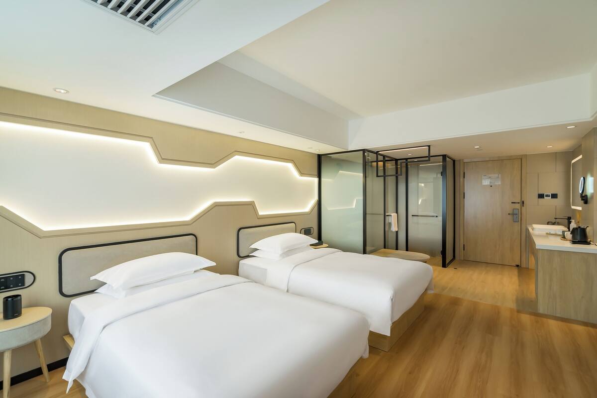 A modern hotel room with two beds, a small desk, a bathroom with a glass partition, and a kitchenette.