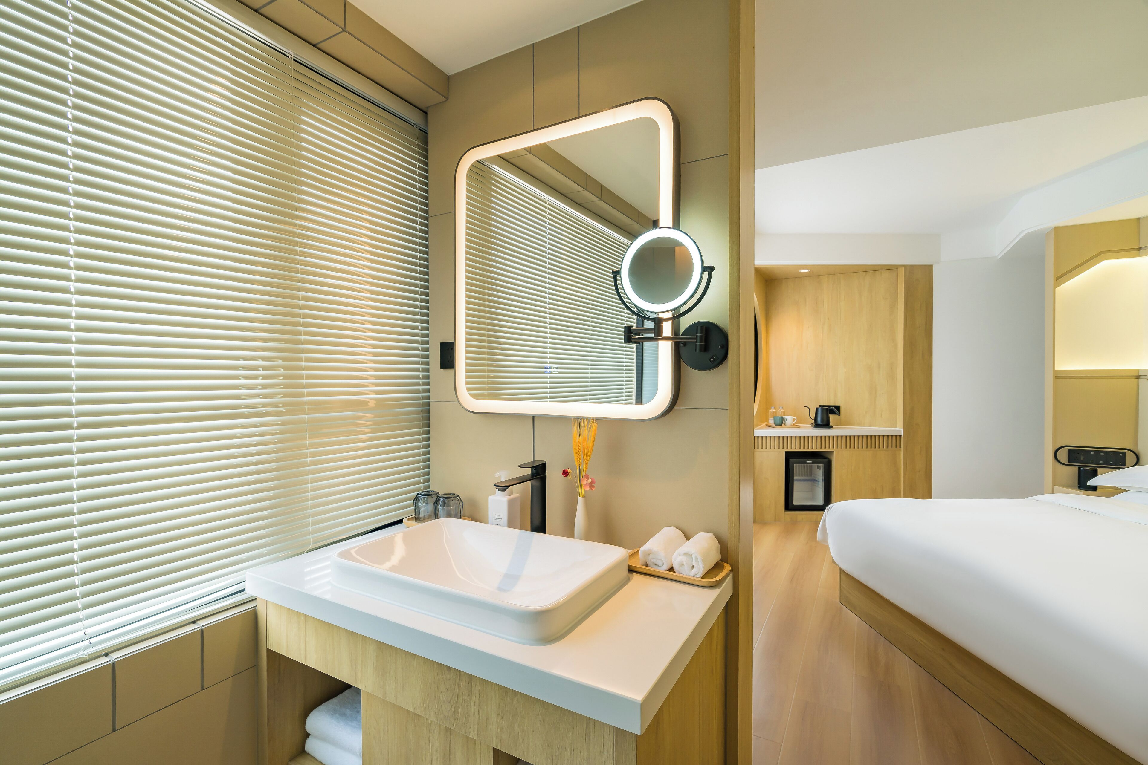 Une salle de bain d’hôtel moderne, dotée d’un grand lavabo, d’un miroir avec éclairage intégré, d’un petit meuble de rangement et d’une table de chevet avec une bouilloire.