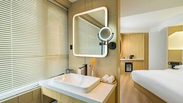 Une salle de bain d’hôtel moderne, dotée d’un grand lavabo, d’un miroir avec éclairage intégré, d’un petit meuble de rangement et d’une table de chevet avec une bouilloire.