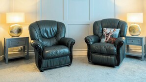 Two black leather armchairs with footrests, positioned on a carpeted floor. Each chair is flanked by a matching side table with a lamp and a decorative sphere. The room features a light blue wall with a framed picture above the armchairs.