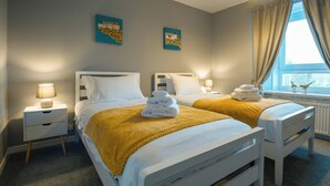 A hotel room with two beds, each with white bedding and a yellow throw blanket. There are two bedside tables with lamps, and artwork on the walls.