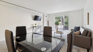 A modern meeting room with a glass table, leather chairs, a small side table, a TV mounted on the wall, and a view of the outdoors through a large window.