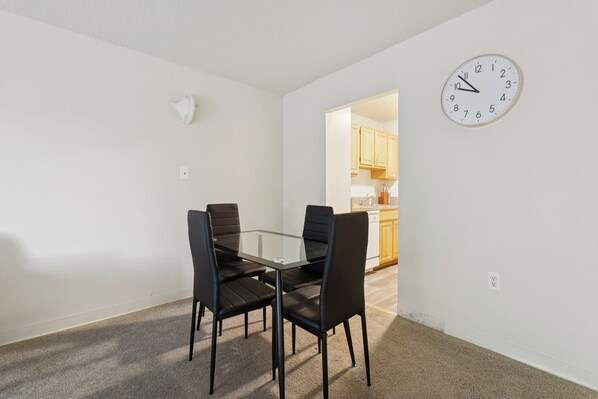 Un comedor con una mesa de vidrio y cuatro sillas, un reloj en la pared y una cocina al fondo.