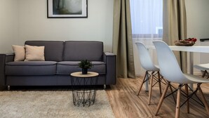Un salon moderne avec un canapé gris, une petite table ronde agrémentée d’une plante, un coin repas avec des chaises blanches et un tableau encadré au mur.