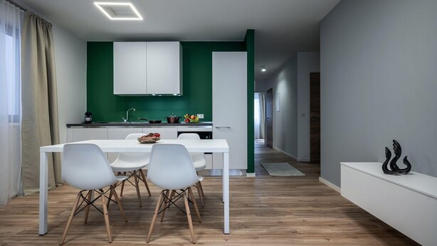 Une cuisine moderne avec des meubles blancs, un mur d’accent vert et un coin repas comprenant une table et des chaises blanches.