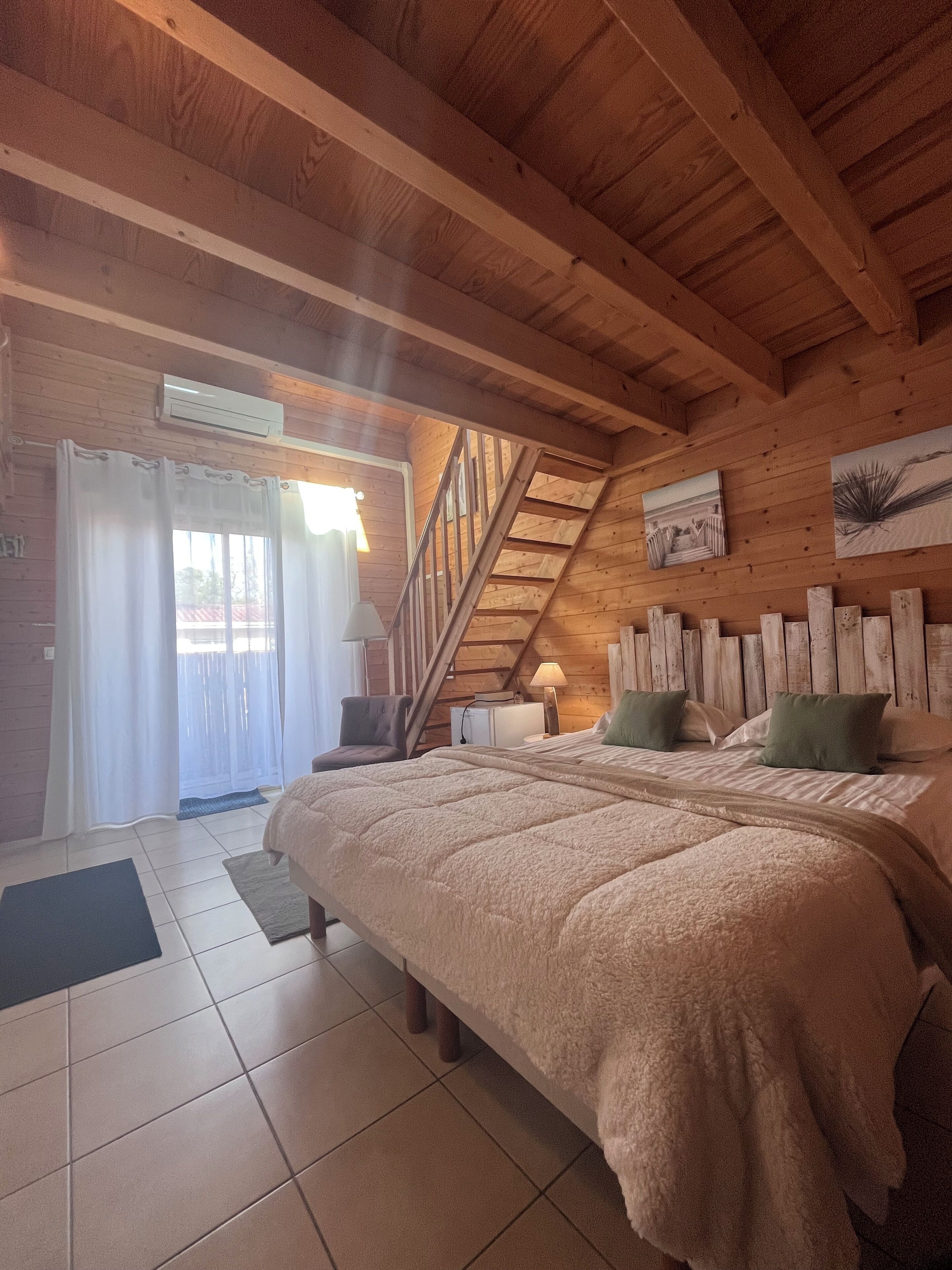 Een slaapkamer met een houten plafond, een bed met een zachte deken, een nachtkastje met een lamp en een raam met gordijnen.
