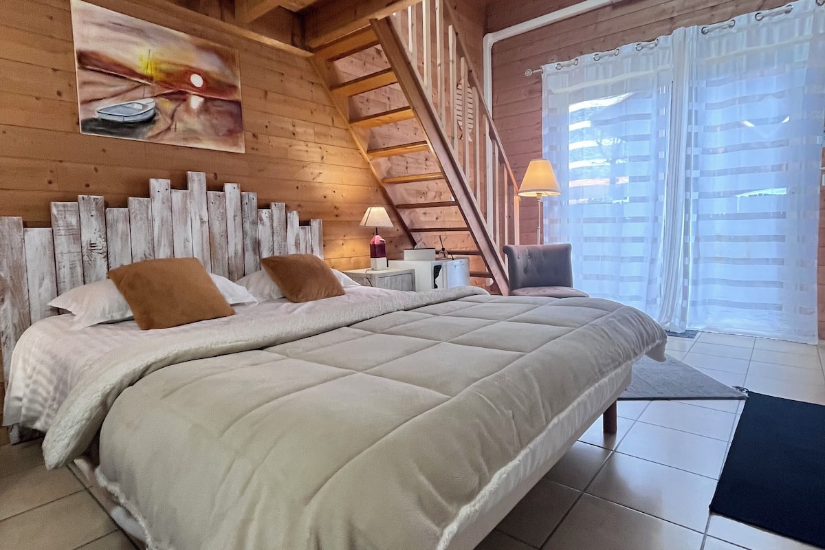 Une chambre à coucher avec un lit, une table de chevet, une lampe, un tableau, un escalier et une fenêtre avec des rideaux.