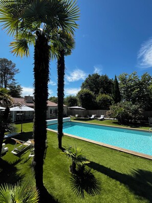 A swimming pool with a wooden deck, surrounded by green grass and palm trees. - VILLA HERBERT (Andernos-les-Bains)