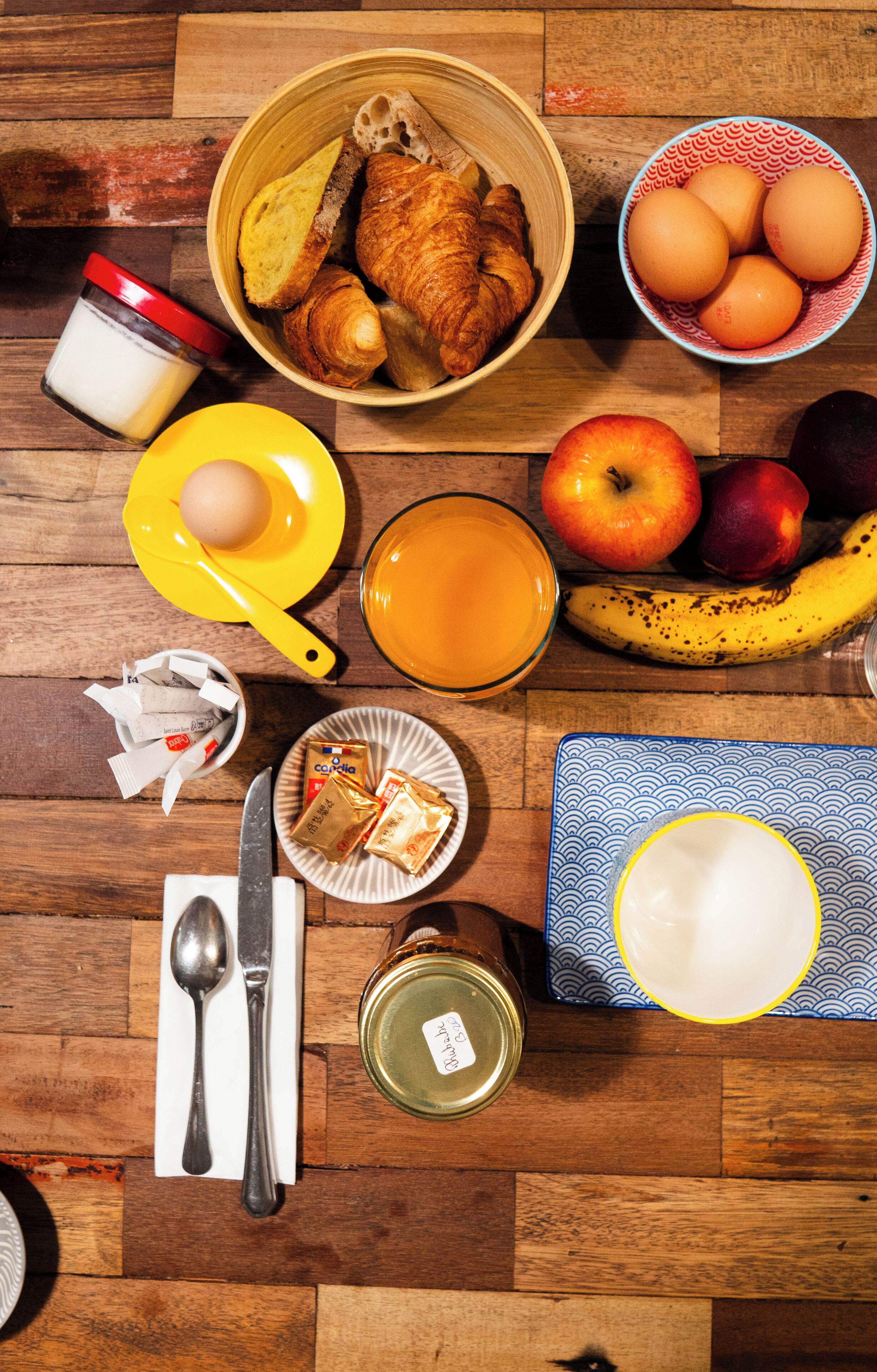 Une table de petit-déjeuner avec des croissants, des œufs, des pommes, une banane, un verre de jus d’orange et divers autres aliments pour le petit-déjeuner.