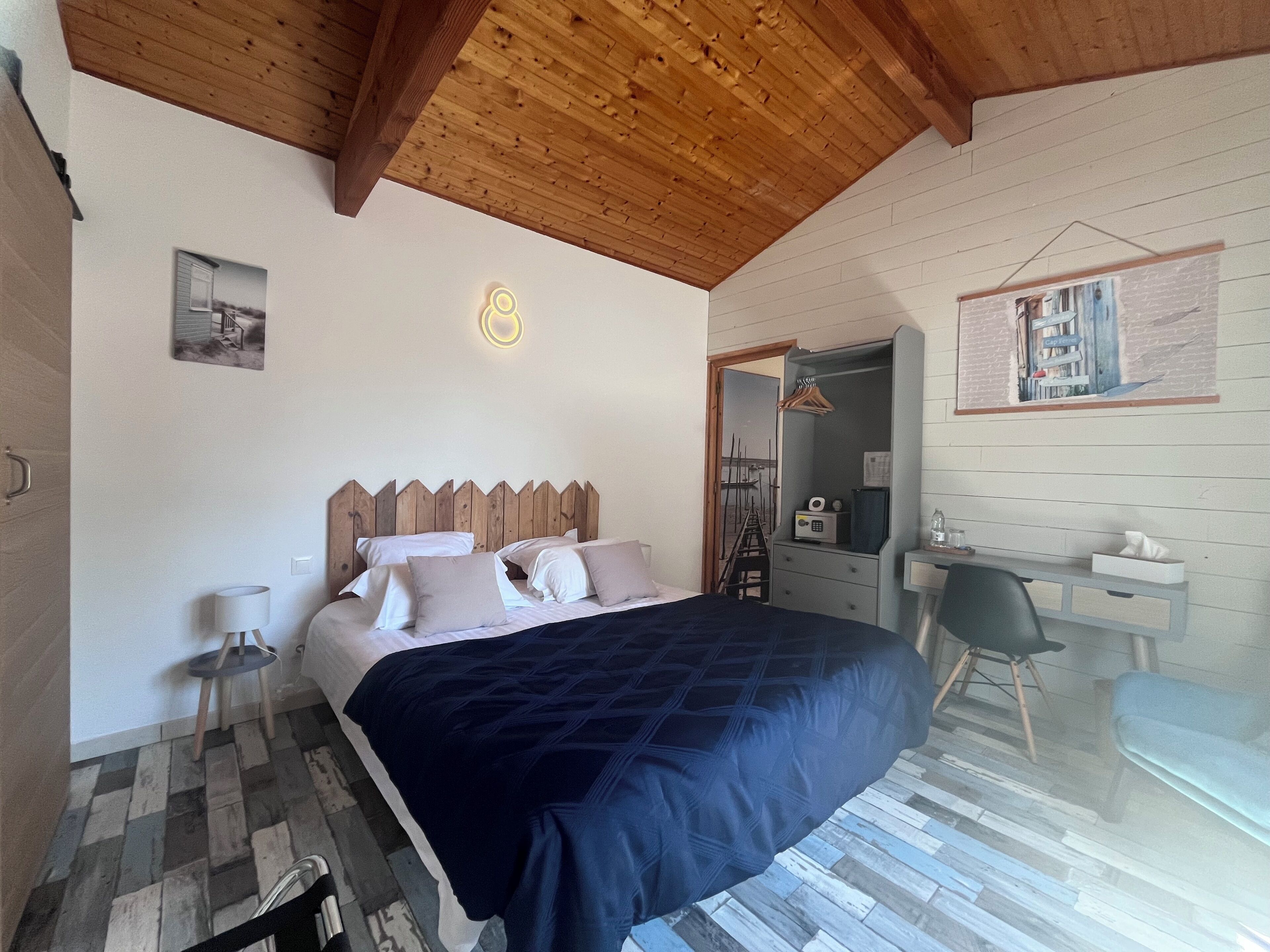 Een slaapkamer met een houten plafond, een bed met donkerblauwe spreideken, een nachtkastje met een lamp, een stoel en een bureau met een spiegel.