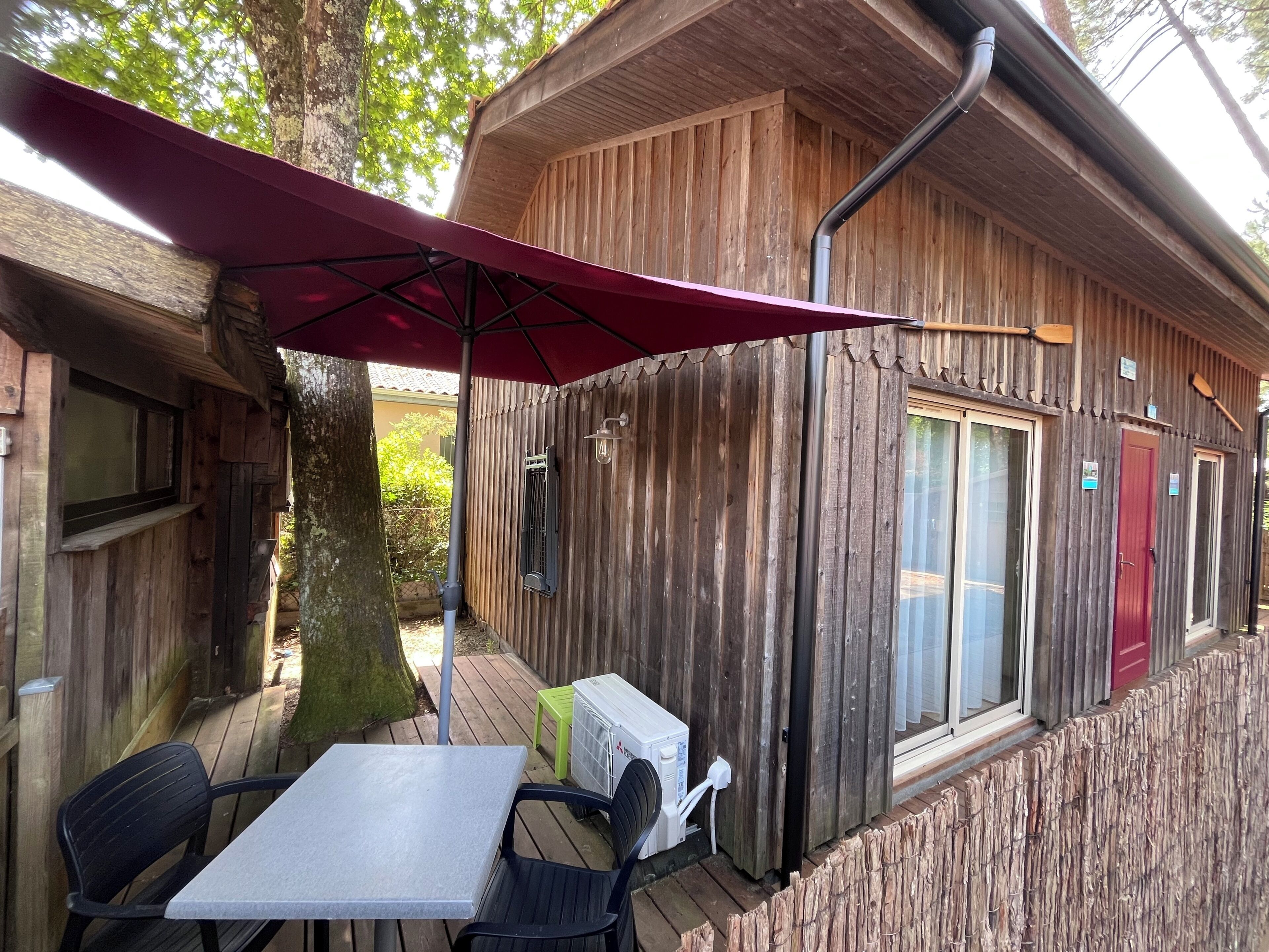 Una cabaña de madera con una mesa y sillas bajo un paraguas rojo, una ventana y una puerta.