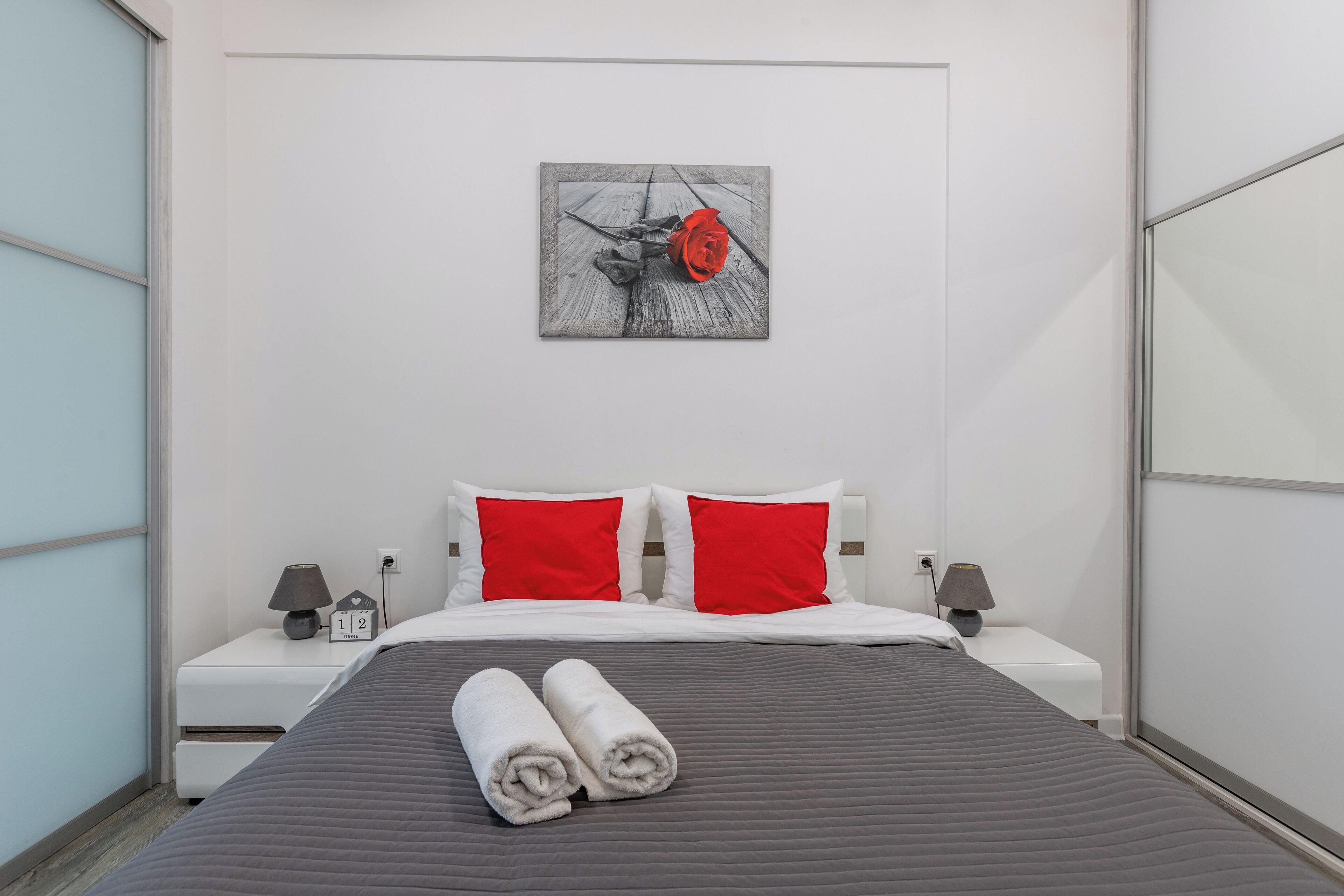 Une chambre d’hôtel moderne avec un lit, deux oreillers rouges, une table de chevet avec une lampe et une horloge murale.