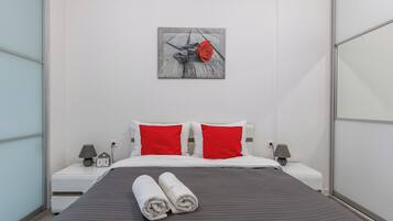 Une chambre d’hôtel moderne avec un lit, deux oreillers rouges, une table de chevet avec une lampe et une horloge murale.