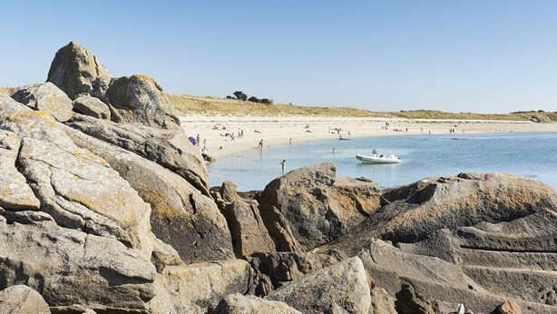 Une plage rocheuse bordée d’une eau d’un bleu limpide, une étendue de sable et un bateau aperçu au loin.