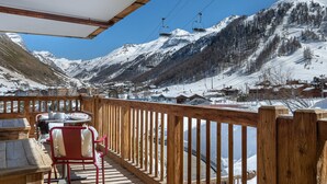 Une terrasse en bois avec une table et des chaises, offrant une vue sur un paysage montagneux enneigé et sur les remontées mécaniques.
