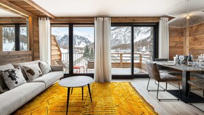 A modern living room with a sofa, a round table, and a dining area with chairs. Large windows offer a view of snowy mountains.