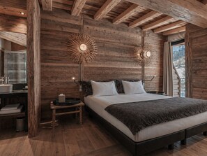 A bedroom with a wooden ceiling, a bed with a dark headboard, two wall-mounted lamps, a bedside table with a lamp, and a window with curtains.
