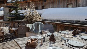 A wooden deck with a table set for dining, snow-covered buildings in the background, and a pine tree in the foreground.