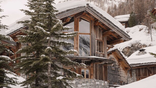 A rustic log cabin with a stone foundation, large windows, and a snow-covered roof.