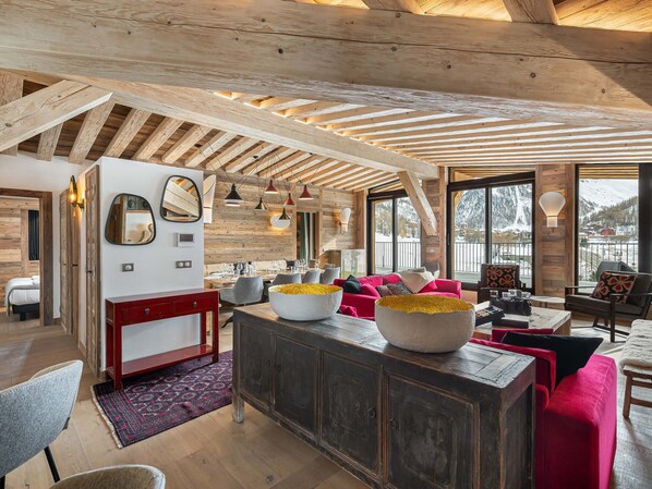 A spacious living area with a wooden ceiling, large windows offering a view of a snowy landscape, a central island with a bowl of flowers, and a red sofa. - Charming Penthouse - 5 Bedrooms - Ski Slope View in Val-d'Isère (Val-d'Isère)