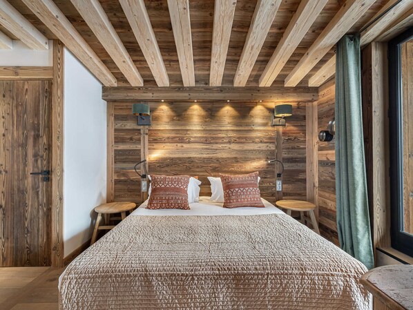 A bedroom with a wooden ceiling, two bedside tables, a bed with patterned bedding, and wall-mounted lamps.