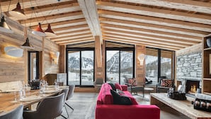 A spacious living area with a wooden ceiling, large windows offering mountain views, a red sofa, and a stone fireplace.