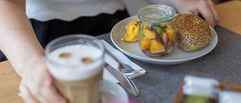 Eine Person hält eine Tasse Kaffee und einen Teller mit einem Sandwich und einem Glas Obstsalat.