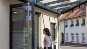 L’entrée d’un hôtel, avec une porte en verre, une enseigne au-dessus indiquant « Hôtel - Pension Treppengasse », et une personne tirant une valise.