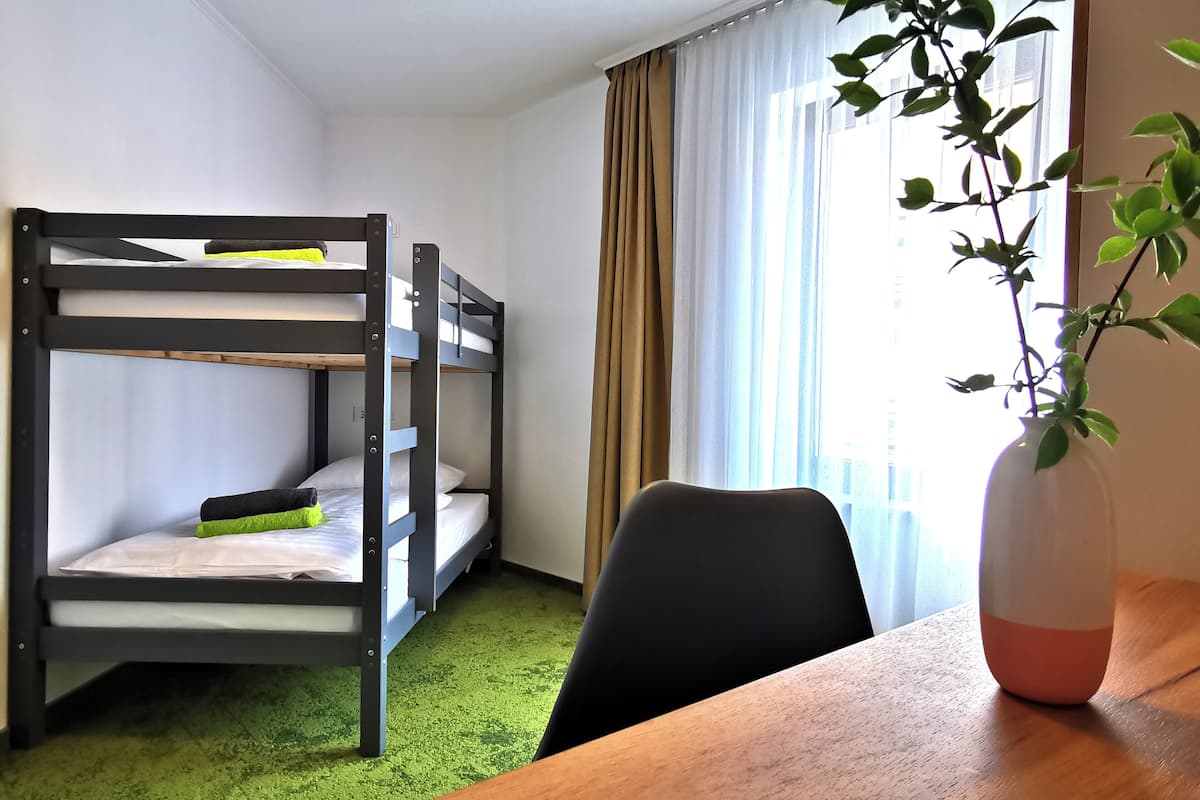 Een slaapkamer met stapelbedden, een groen tapijt, een houten tafel, een zwarte stoel en een potplant.