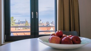 Une pièce donnant sur un paysage urbain, avec une fenêtre à cadre noir et une table blanche sur laquelle se trouve un bol de pommes rouges.