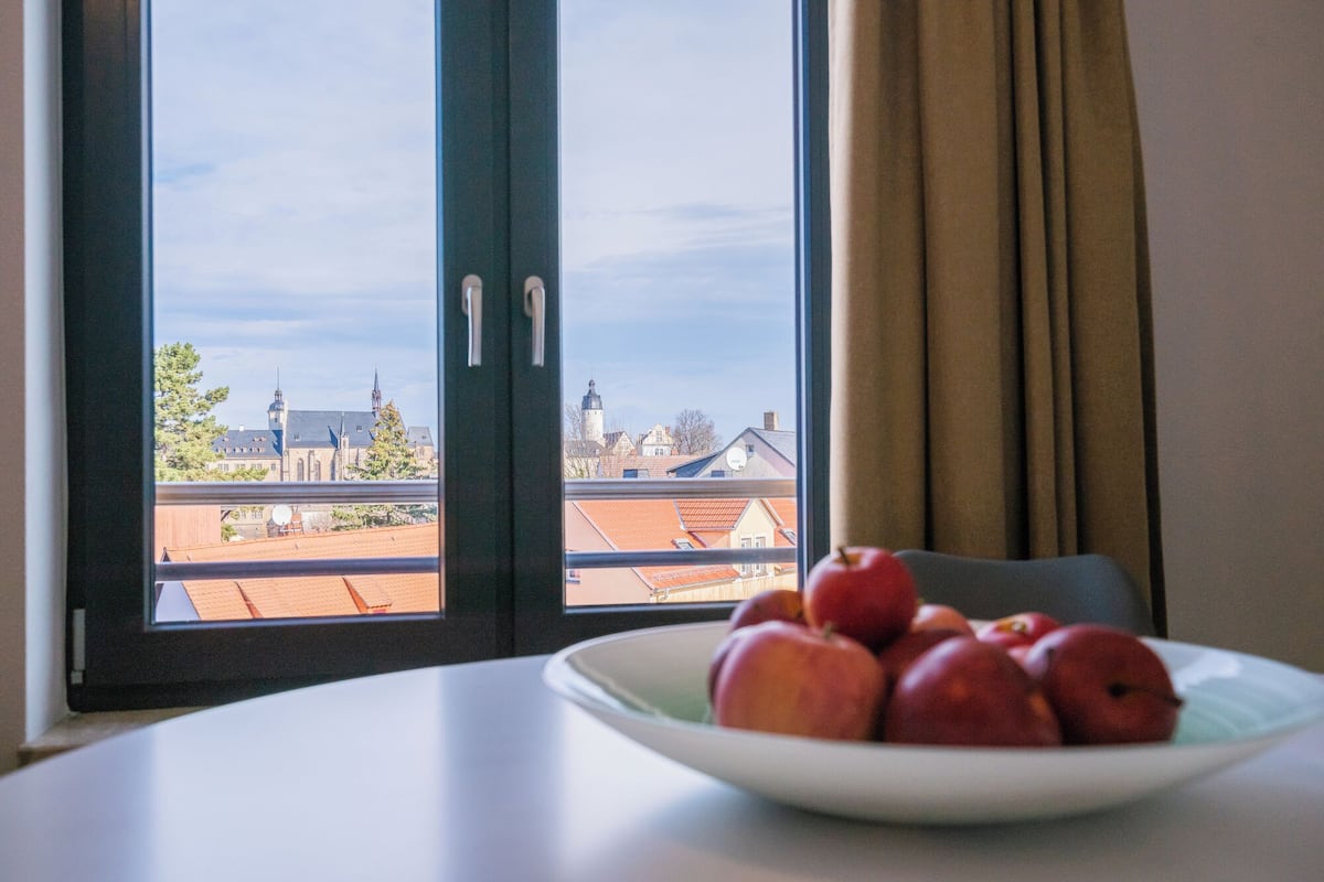 Een kamer met uitzicht op een stadsgezicht, door een raam met zwarte kaders. Er staat een witte tafel met een schaal rode appels.