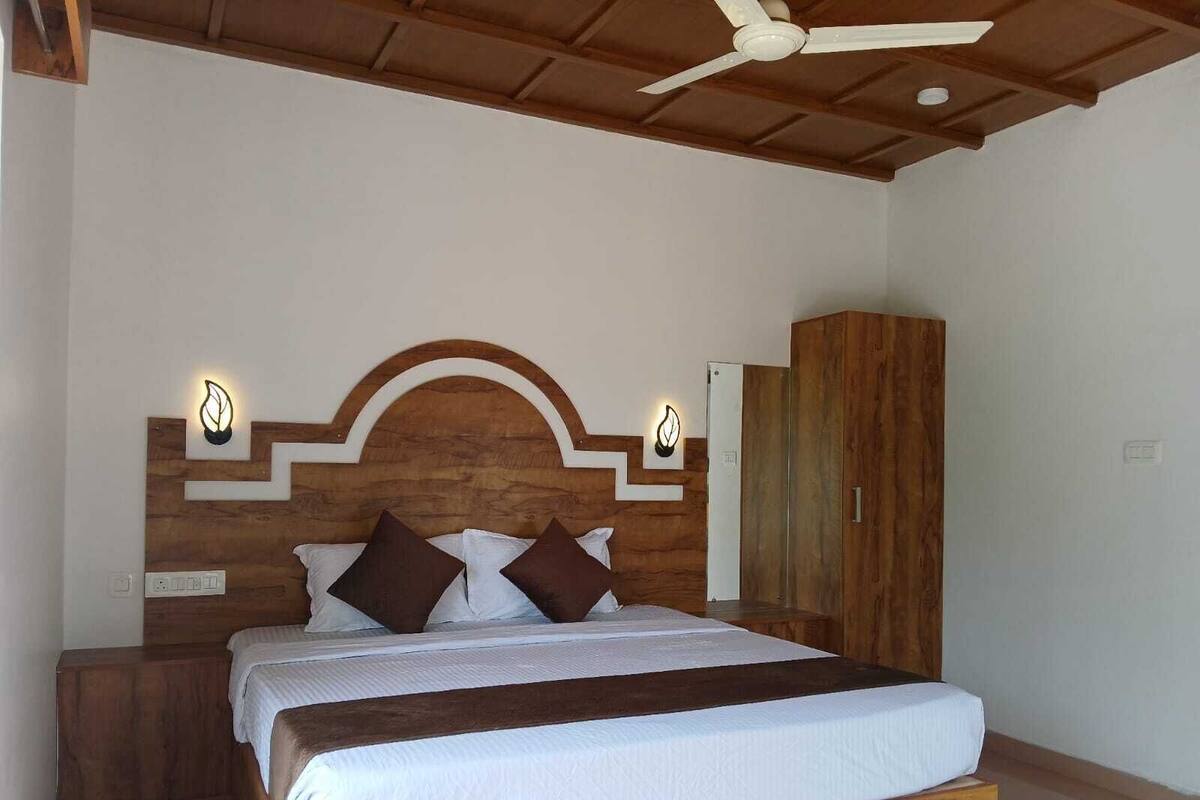 A bedroom with a wooden headboard, a bed with white bedding and brown pillows, a nightstand, a wardrobe, and a ceiling fan.