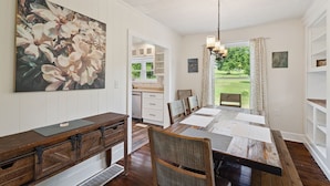 A dining area with a wooden table and chairs, a painting of flowers on the wall, and a view of a garden through a window. - A peaceful retreat just off the Seneca Lake Wine Trail—close to 20+ wineries and Smith Park’s lakeside fun. (Hector)
