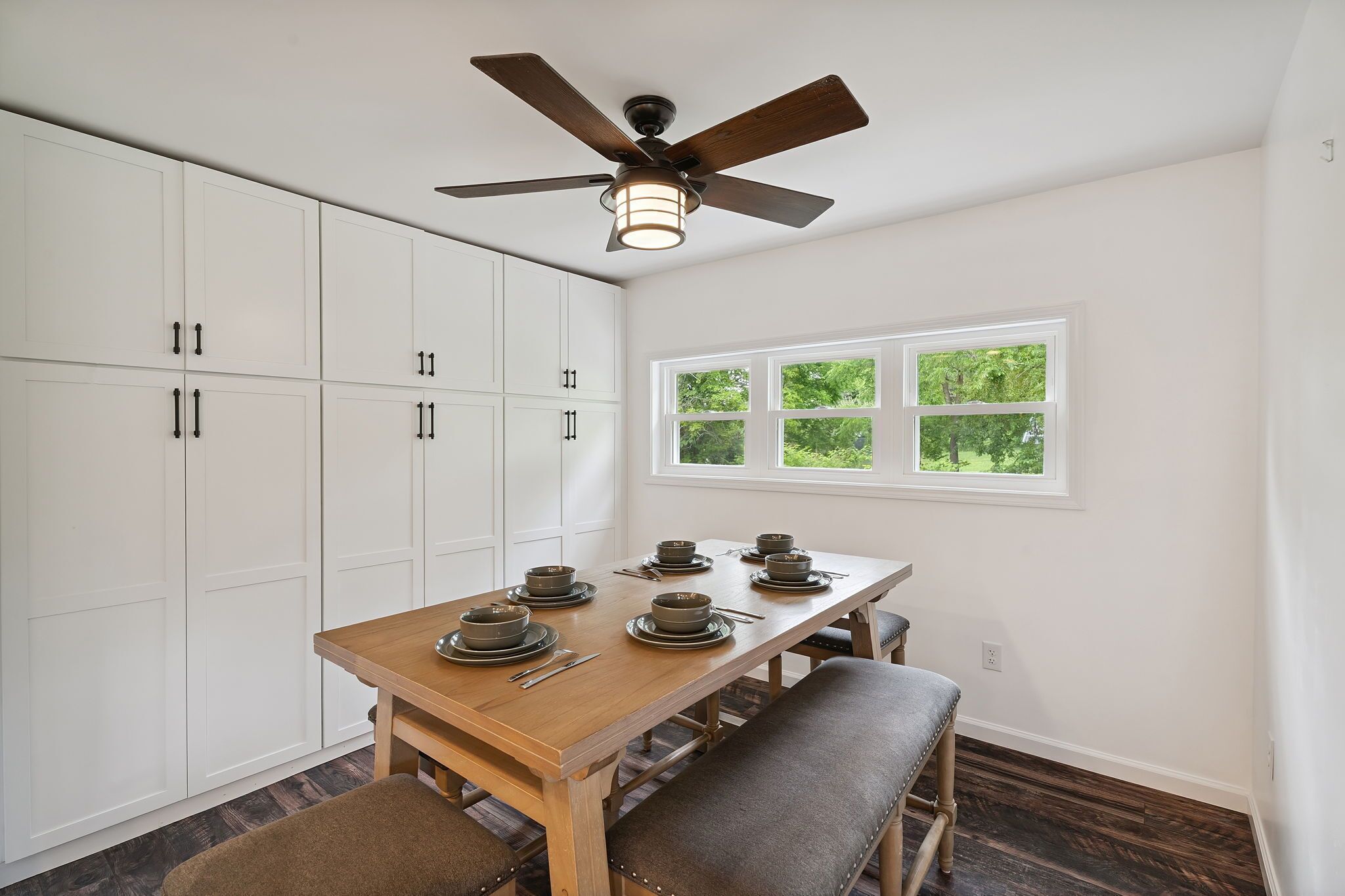 A dining area with a wooden table set for six, white cabinets, and a ceiling fan.
