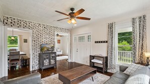 A spacious living room with a brick accent wall, a ceiling fan, a wooden coffee table, and a view of the outdoors through a window with patterned curtains. - A peaceful retreat just off the Seneca Lake Wine Trail—close to 20+ wineries and Smith Park’s lakeside fun. (Hector)