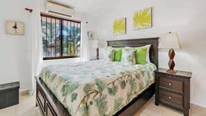 A bedroom with a bed, a nightstand, a lamp, and a window with a view of palm trees. - Green Turtle 20 in Coco is an elegantly appointed 2-bedroom, 1-bath condo with a swimming pool located within a 7-minute walk of the beach. <br><br>📢Important announcement: Golf cart with condo available upon request for additional charges (Playas Del Coco)