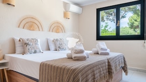 Een slaapkamer met een groot bed, twee wandventilatoren, een raam met uitzicht op bomen en een wandairconditioner.