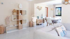 Een moderne woonkamer met een bank, een eettafel met stoelen en een keukenhoek met een wit kastje en planken.