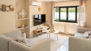 Een moderne woonkamer met een zithoek, een televisie, een tafeltje en een raam met uitzicht op groen.