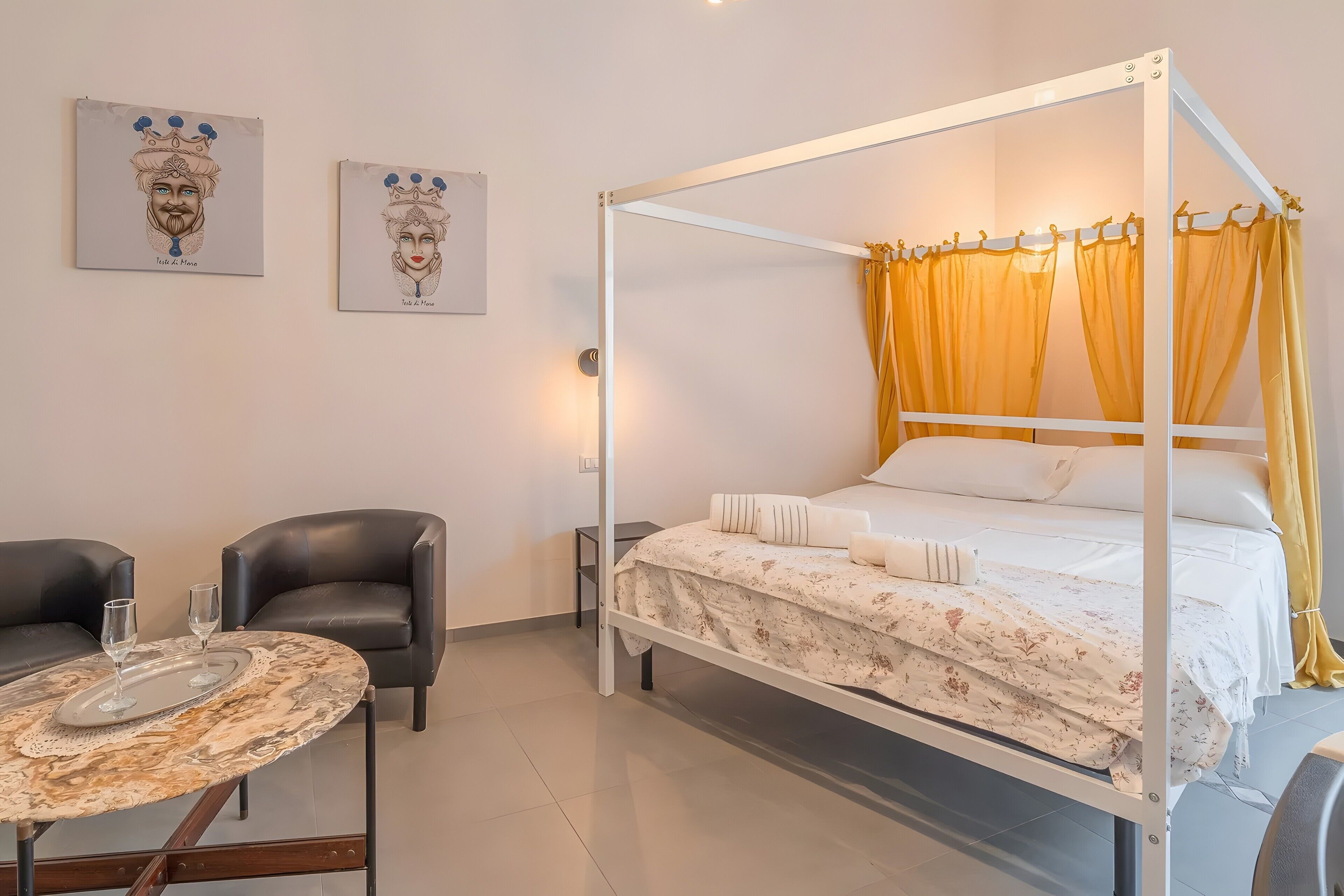 Una cama con dosel, cortinas amarillas, una mesita de mármol con vasos, dos sillas negras y dos cuadros en la pared.