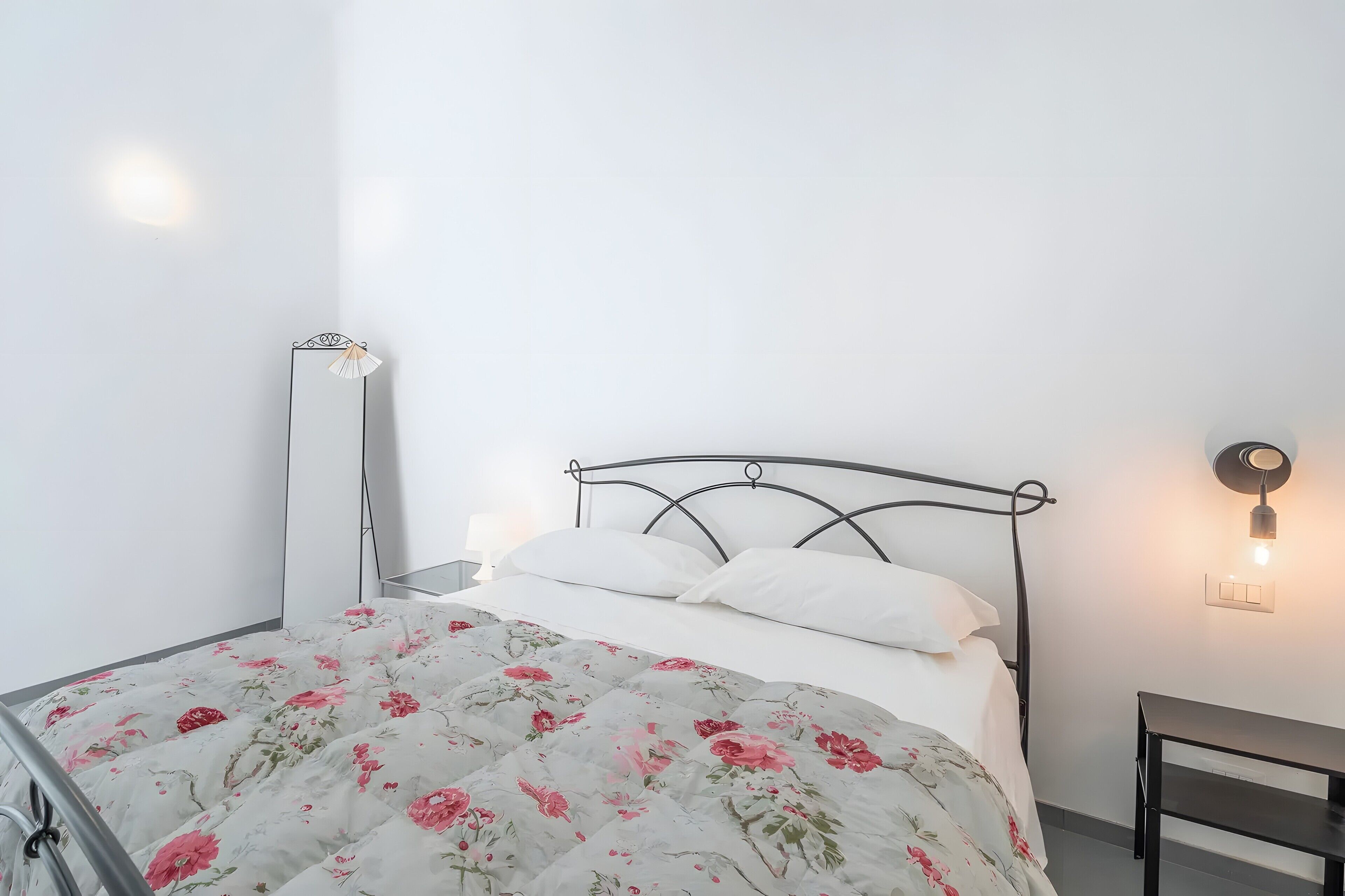Una cama con colcha estampada, una mesita de noche, una lámpara de pie y un aplique de pared.