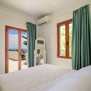 Un dormitorio con cama, ventana con cortinas, puerta que da a un balcón exterior y un equipo de aire acondicionado en la pared.